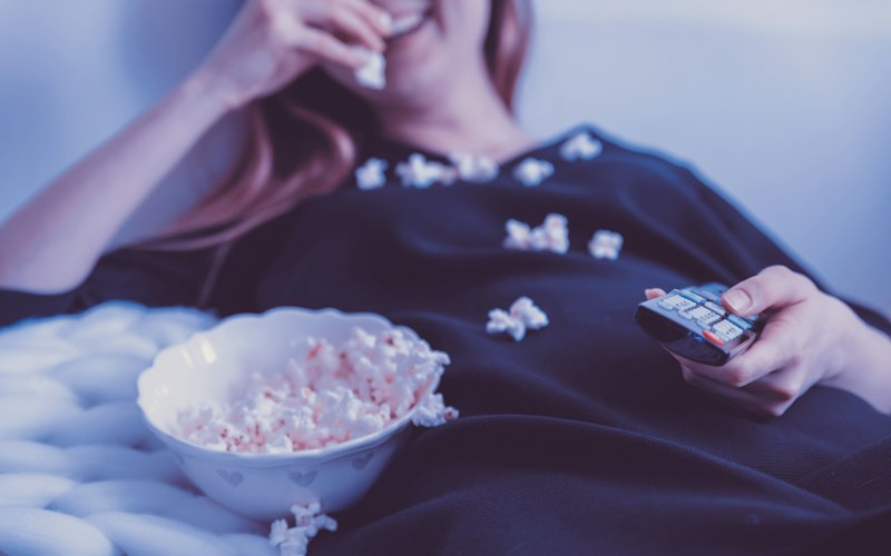 A woman curled up under a blanket with a bowl of popcorn and a remote