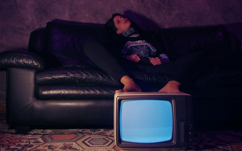 A woman slumped on a dark leather sofa, feet up behind a glowing retro TV
